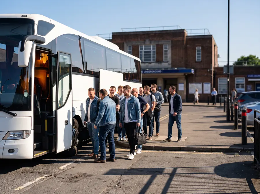 A modern white touring coach parked at the kerb on the forecourt outside Stanmore Underground station on a bright clear summer morning, its door open and interior lit, a group of men in their twenties and thirties in casual clothes boarding for a day out in front of the 1930s brick station building.